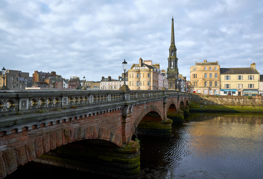 Stone arch bridge spanning a river with historic buildings and church spire beyond in soft daylight.