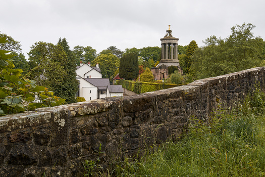 Stone wall in foreground frames rural landscape with white cottage and bell tower in middle distance surrounded by dense green trees.