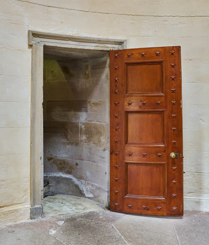 Wooden door with decorative studs open to reveal a concrete interior room with white walls.