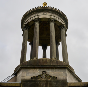 Neoclassical cylindrical monument with fluted columns and decorative cornice against overcast sky.