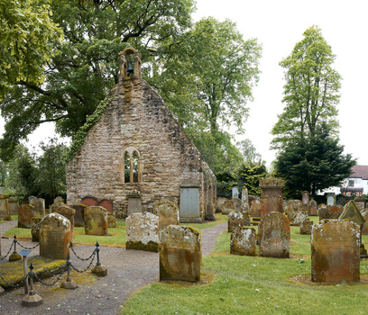 Stone chapel surrounded by weathered gravestones in a cemetery beneath mature trees.