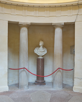 Neoclassical bust on a pedestal framed by fluted columns and red velvet ropes in a gallery alcove.