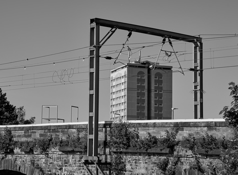 Black and white photograph of electrical transmission equipment silhouetted against modern apartment buildings.