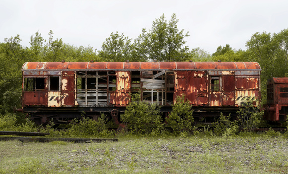 Abandoned red railroad car with broken windows surrounded by overgrown vegetation and trees.