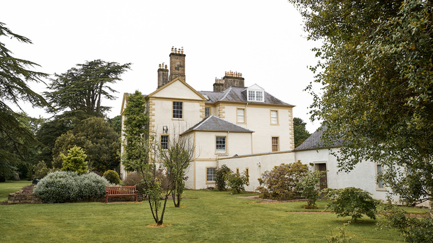 Cream-colored country manor house with multiple chimneys surrounded by manicured gardens and mature trees.