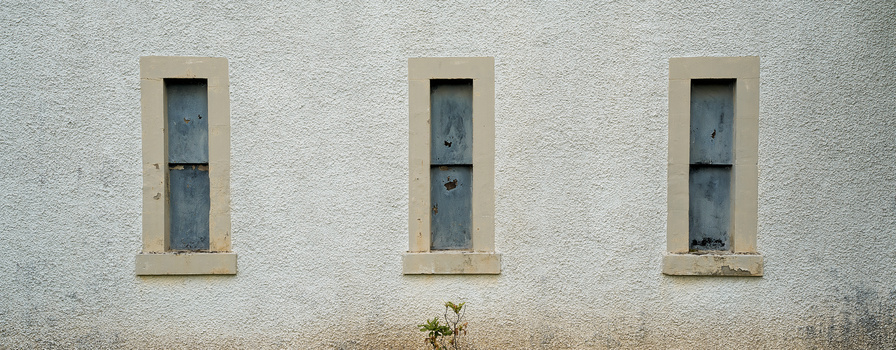 Three vertical rectangular windows with weathered blue-gray panels set in a cream-colored concrete wall.