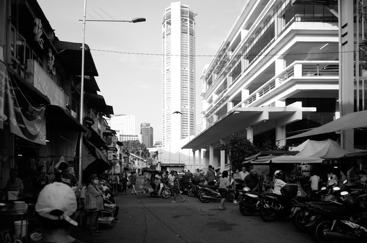 Busy urban street scene with motorcycles, pedestrians, and market stalls beneath modern high-rise buildings.