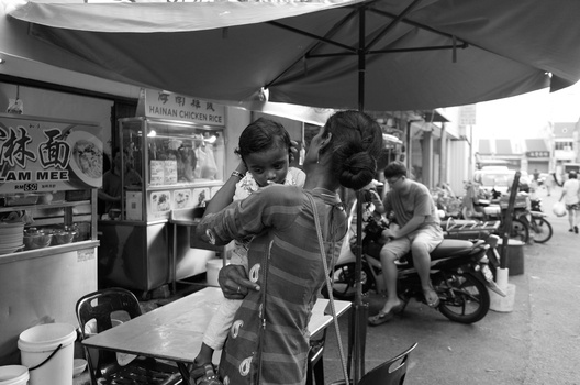 Two people conversing under a large umbrella at a street market stall in black and white.