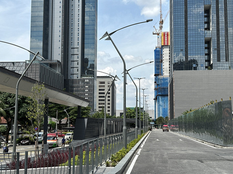 Urban street with modern curved street lamps, high-rise buildings, and green vegetation barriers under partly cloudy sky.