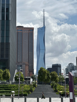 Modern urban plaza with distinctive curved blue tower framed between classical and contemporary buildings under dramatic cloudy sky.