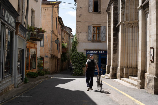 Person on bicycle riding through a narrow European street lined with ochre and stone buildings.