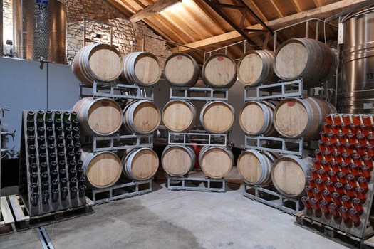 Oak barrels stacked in three horizontal rows inside a winery facility with exposed brick and steel support structures.