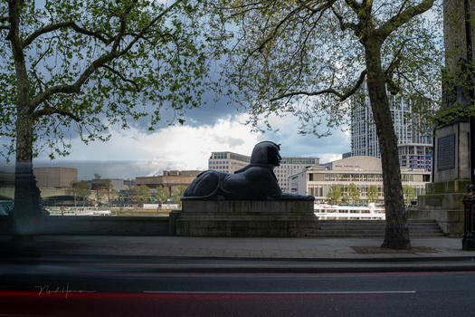 Black reclining figure sculpture on a waterfront promenade with city skyline beyond.