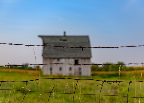 Barbed wire fence in sharp focus with a blurred abandoned house in the background across a green field.
