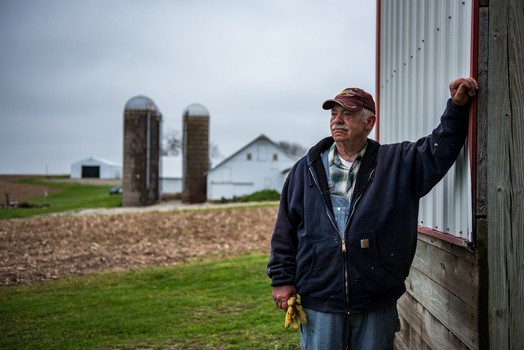 Farmer in navy jacket and cap stands beside corrugated metal barn wall, holding it with one hand, with silos and farmhouse visible across green fields.