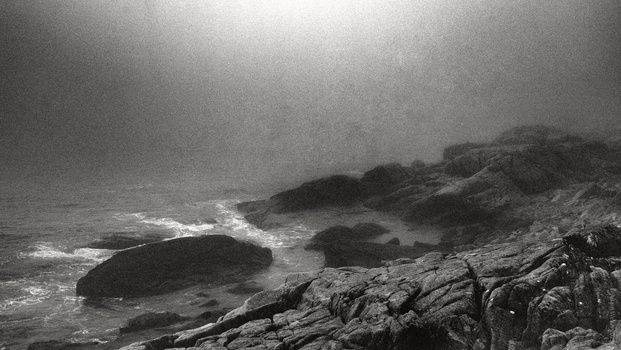 Dramatic black and white coastal landscape with rocky outcrops and turbulent ocean under overcast sky.