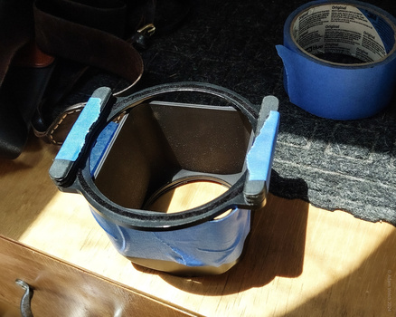 Metal bucket with blue handles containing white paint and a brush, positioned in natural sunlight on a wooden surface.