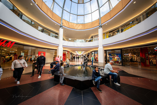 Modern shopping mall interior with circular skylight, central fountain, and multiple retail levels bathed in warm ambient lighting.