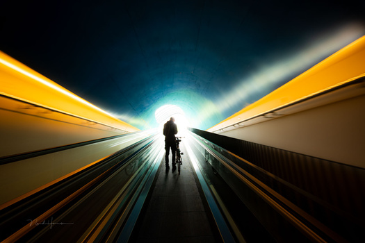 Silhouetted figure with bicycle standing in a tunnel, backlit by bright light ahead.