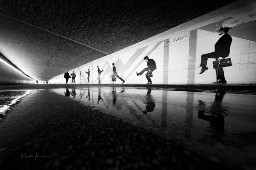 Silhouetted figures skateboarding across a wet, reflective underground passage with dramatic overhead lighting.