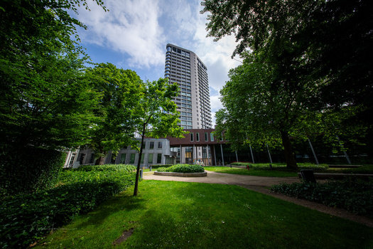 Modern high-rise residential tower framed by mature green trees and a low-rise building in a park setting.