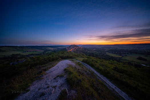 Rocky ridge in foreground overlooking illuminated town nestled in valley at twilight.