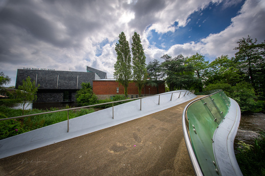 Modern curved pedestrian bridge with metal railings spanning over a waterway, surrounded by green vegetation and contemporary architecture.
