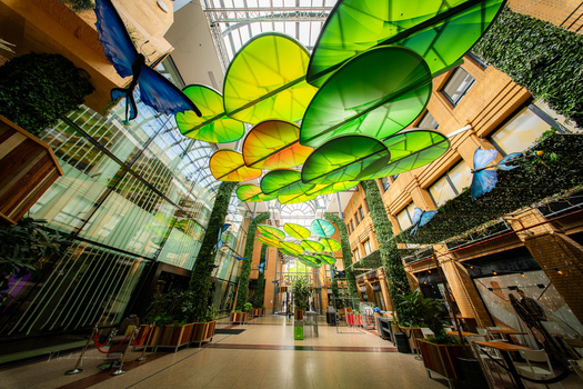 Vibrant green and yellow sculptural orbs suspended from a glass ceiling in a modern architectural corridor.