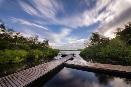 Wooden boardwalks intersecting over still water with mangrove trees lining both sides under a dramatic cloudy sky.