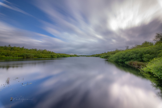 Tranquil river landscape with lush green vegetation lining both banks and dramatic clouds reflected in still water.