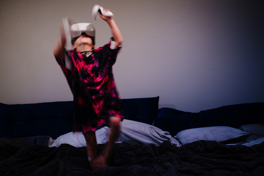 Child in red patterned shirt on bed holding up VR headset with both hands.