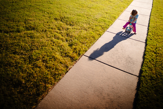 Child riding a scooter on a concrete pathway with manicured lawn on both sides, casting a long shadow in bright sunlight.