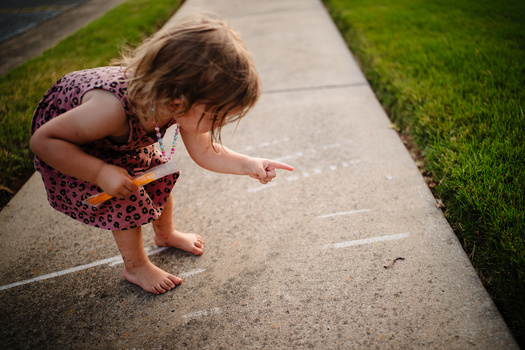 Young child crouching on concrete sidewalk, drawing with chalk.