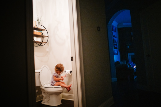 Young child sitting on toilet in brightly lit bathroom doorway, framed by dark hallway.