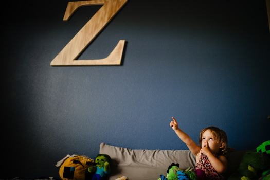 Toddler pointing upward at large wooden letter Z mounted on blue wall above toys.