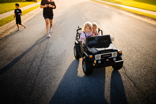 Two children in a toy electric vehicle driving on a suburban road while two joggers run alongside.