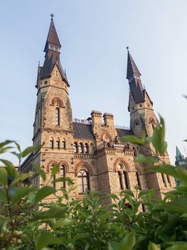 Gothic Revival brick church with twin spires framed by green foliage in foreground.