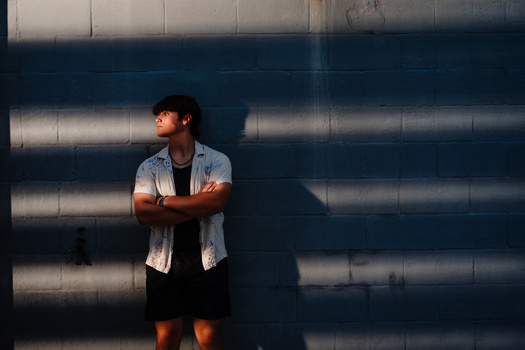 Man in light shirt standing against industrial metal wall with dramatic shadow patterns.