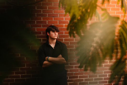 Young man in dark clothing standing against a red brick wall, backlit by warm golden light filtering through leafy foliage.