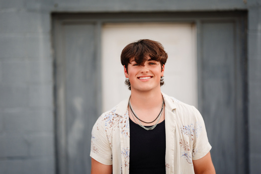 Young man smiling at camera wearing black v-neck shirt and cream button-up cardigan with floral pattern.