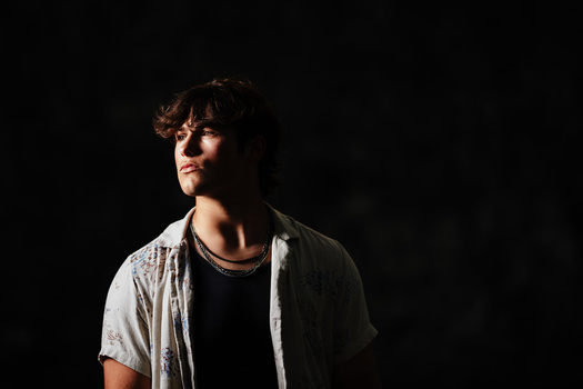 Young man with dark wavy hair wearing glasses, a black undershirt, and light gray button-up shirt, photographed against a dark background.