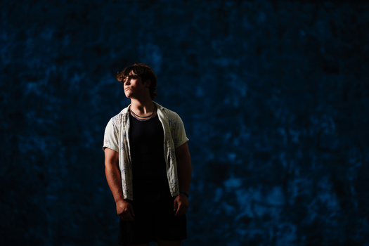 Young man in casual shirt standing against textured blue wall, looking to the side.