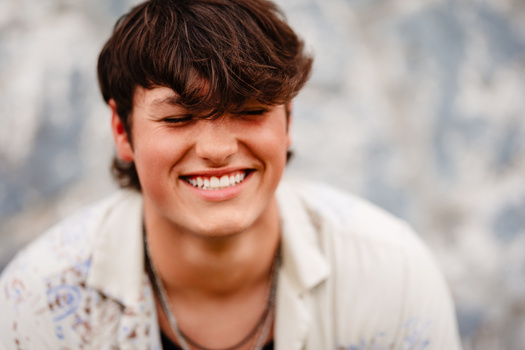 Young man with dark hair smiling at camera against blurred gray and white background.