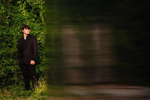 Man in dark suit standing against lush green ivy wall with blurred background.