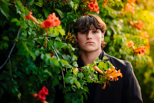 Young person with dark hair surrounded by vibrant orange and red flowering vines, wearing black clothing and gazing to the side.