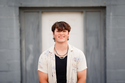 Young man with dark hair smiling at camera wearing white patterned shirt over black tee.