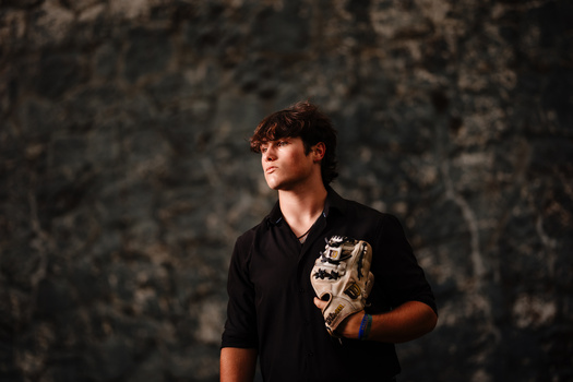 Young man in black shirt holding a baseball glove against textured stone wall.