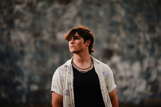 Young man with reddish-brown hair wearing sunglasses, black shirt, and light patterned overshirt, gazing upward against a textured concrete wall.