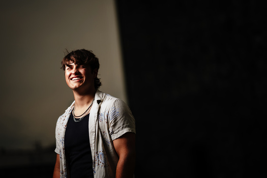 Young man in casual shirt smiling against dramatic lighting with strong shadow.