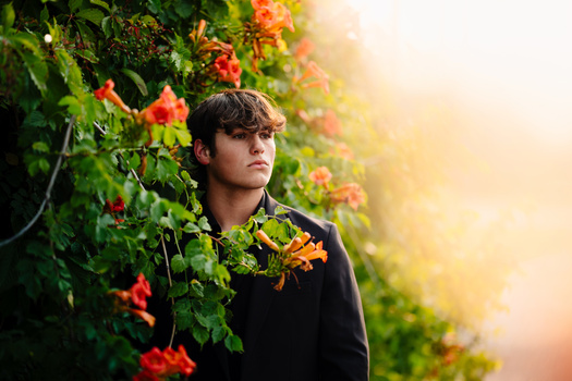 Young man in dark jacket standing among vibrant orange and red flowering vines during golden hour.
